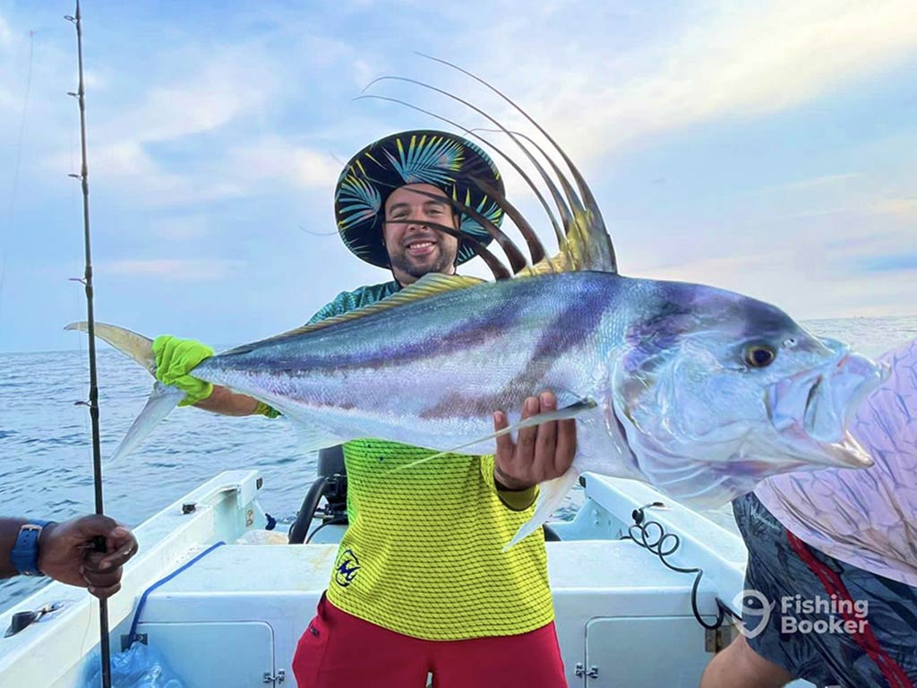 A smiling angler in a yellow shirt and big hat struggles to hold a large Roosterfish with the water behind him on a cloudy day