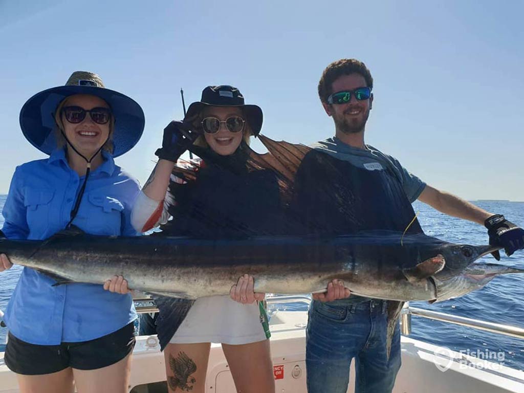 Three anglers on a fishing charter in the Northern Territory – two female, one male – holding a large Sailfish on a sunny day with no clouds in the sky