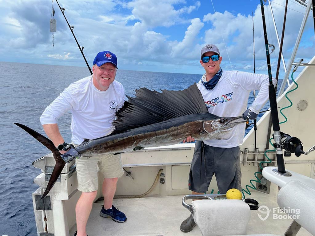 Two anglers in white shirts and baseball caps, hold a large Sailfish aboard a fishing charter in the Florida Keys, with the water behind them on a day with sunny intervals