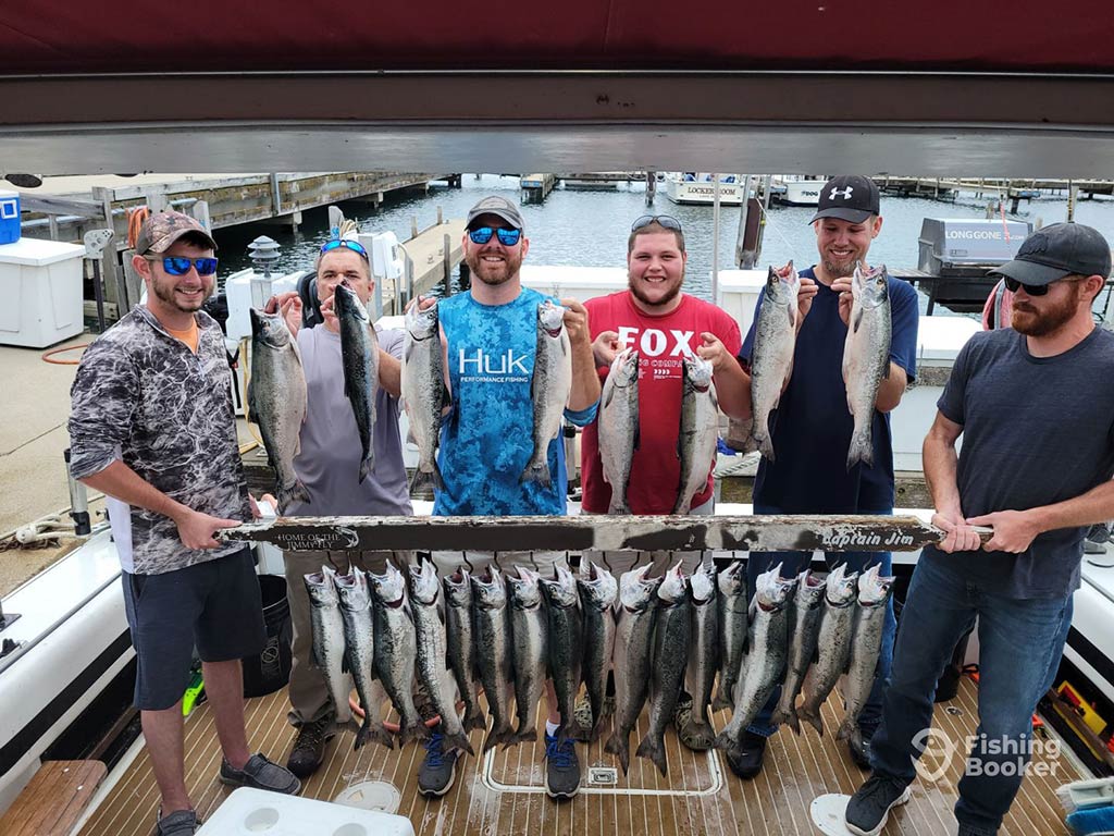 A group of anglers stand on the deck of a fishing boat, holding up a number of Salmon, with a dozen or so more hooked from a wooden sign in front of them