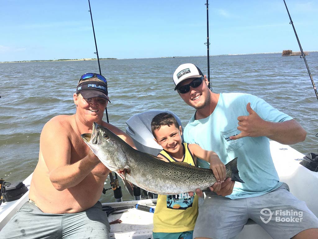 Two men pose either side of a young boy, while holding a large Spotted Seatrout aboard a fishing charter in Manteo, with the water and two fishing rods behind them on a clear day