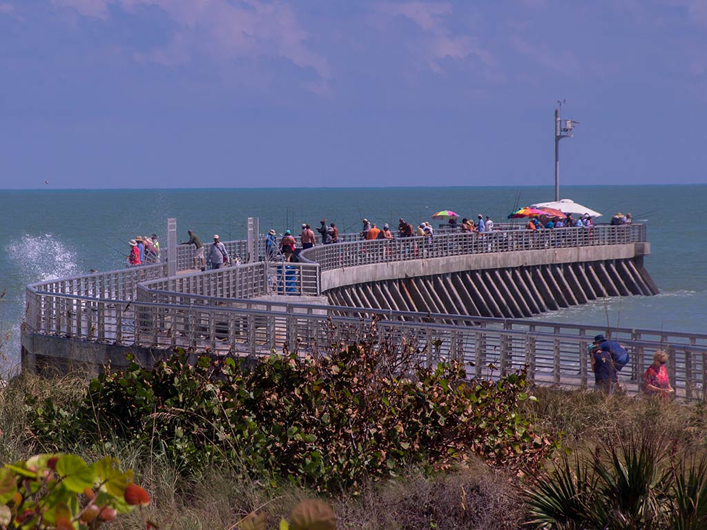 A view from a bank towards one of the fishing jetties in Sebastian, FL, full of people on a clear day, with the ocean visible in the distance