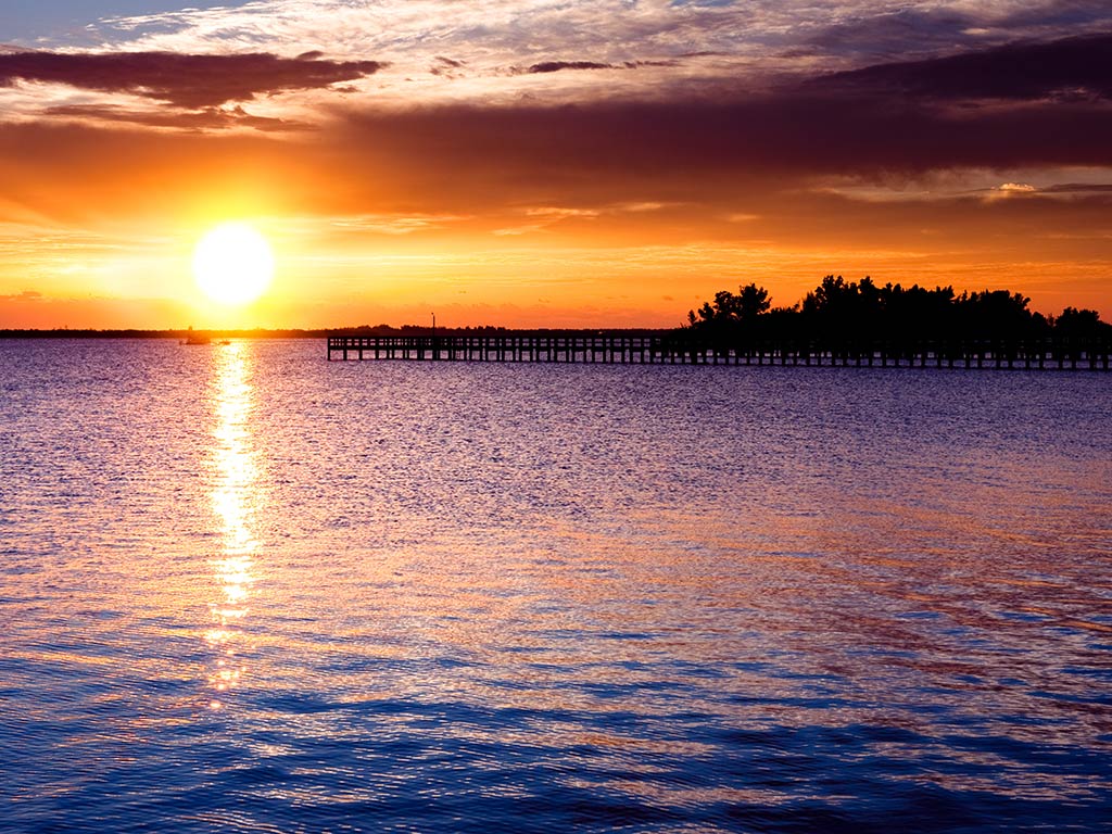 A view across the water towards a fishing pier in Sebastian, FL at sunset, with the sun setting in the distance, creating an orange hue in the air