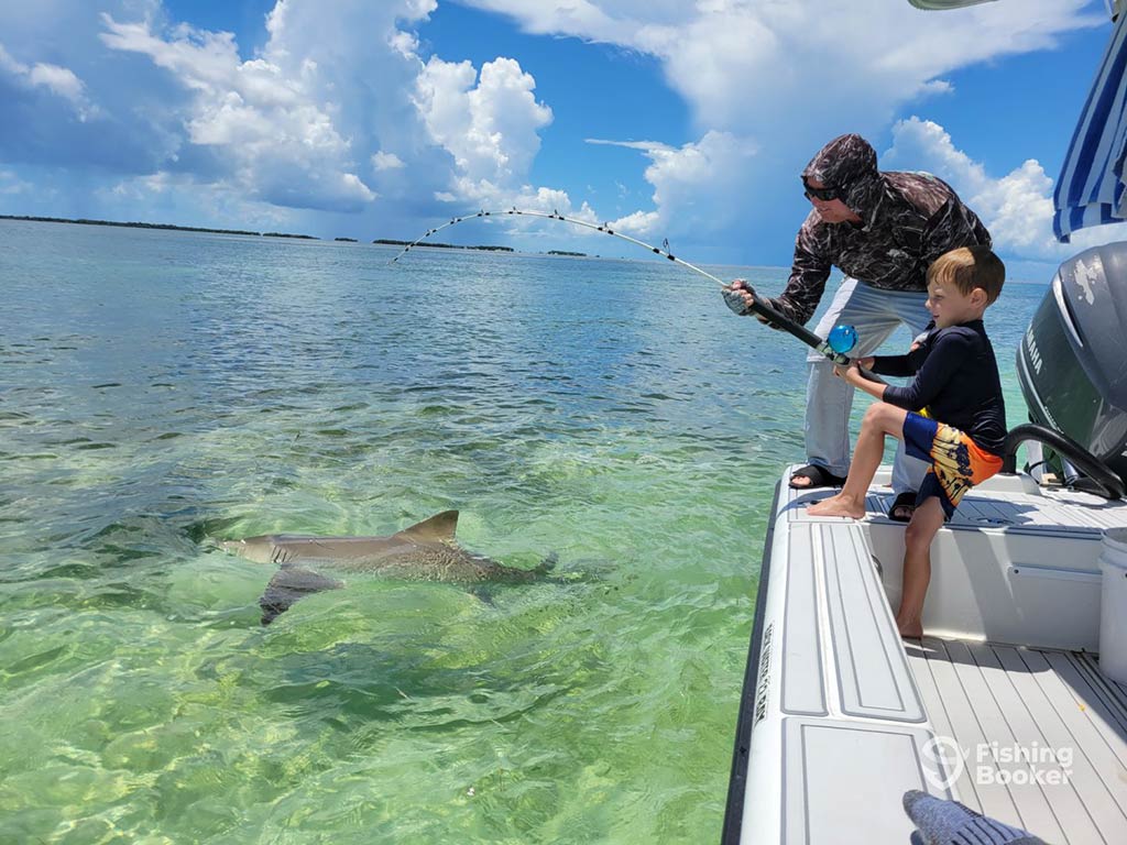 A small boy steadies himself on the side of a fishing boat, as he attempts to reel in a Shark, visible in the clear waters, while a man helps him on a sunny day in the Florida Keys