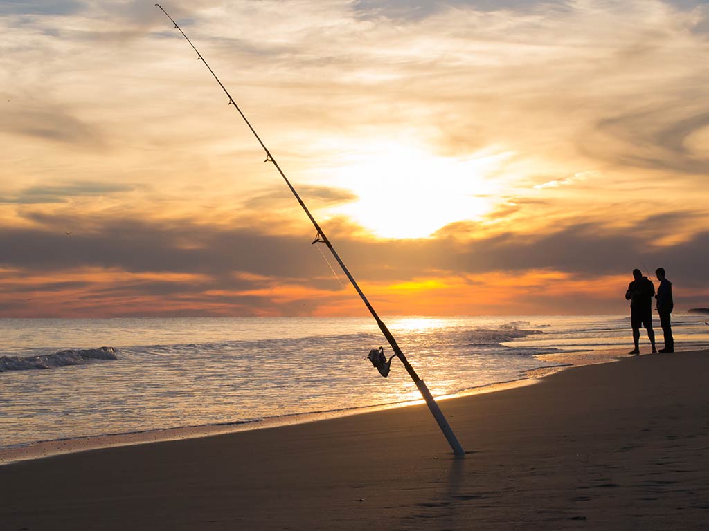 A lone fishing rod stands in its holder on a beach in the Outer Banks at sunset, while two people stand as silhouettes in the distance on the right