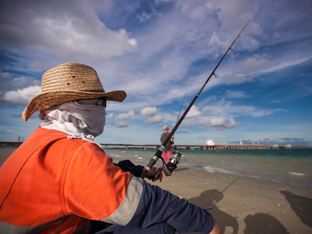 A closeup of the side of an angler in a red t-shirt, face bluff and a straw hat, holding a fishing rod and casting into the surf, as he crouches on a beach on a sunny day