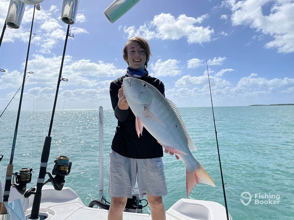 A teenage boy stands on a fishing charter, holding a large Snapper, caught near Stock Island on a clear day, with turquoise waters and blue skies with white clouds behind him