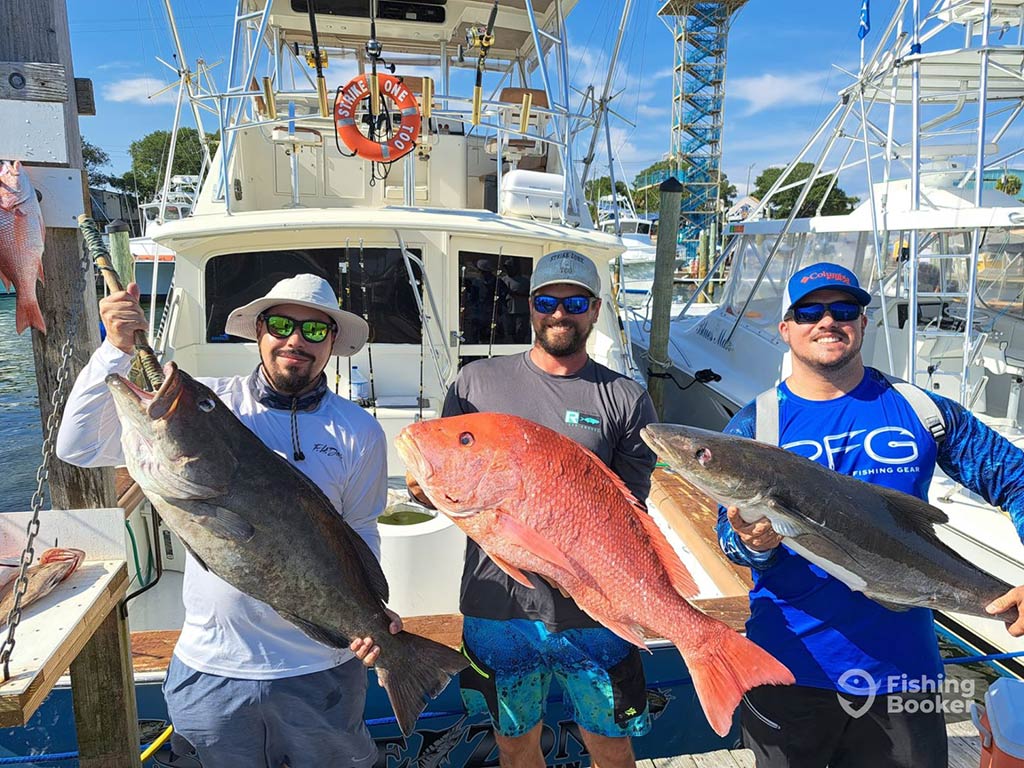 Three anglers stood on the back of an offshore fishing charter, the one in the middle holding a Red Snapper, the one on the right holding a Grouper, and the one on the left of the image holding a Cobia on a sunny day in the Florida Panhandle