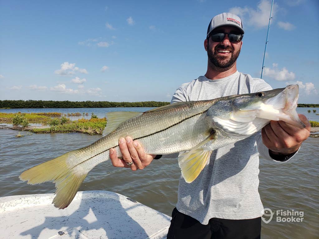 A smiling angler in a baseball cap and sunglasses standing on a fishing boat in Port Isabel, TX, holding a large Snook, with the water and mangroves visible behind him on a sunny day