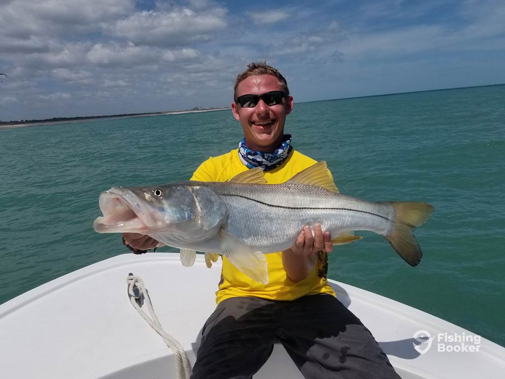 An angler wearing sunglasses and a yellow shirt, sits on the front of a fishing charter in Sebastian, holding a large Snook, with the water behind him on a sunny day with some clouds