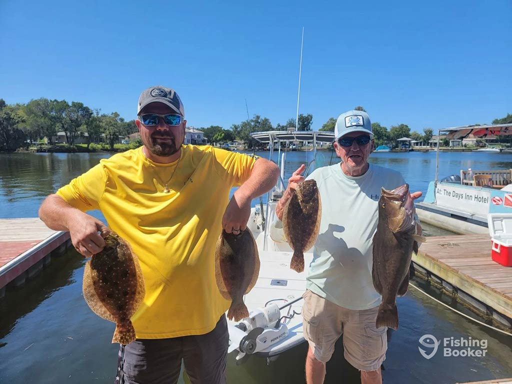 Two anglers proudly pose in front of a fishing boat at a dock in Florida, holding three Flounder and one other fish between them on a sunny day