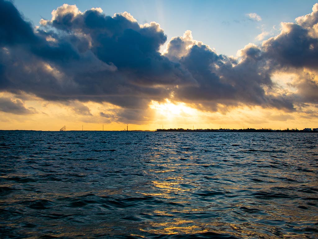 A view across the blue water out of Stock Island, FL, at sunset, with the sun setting over some distant land and a few clouds creating an impressive image