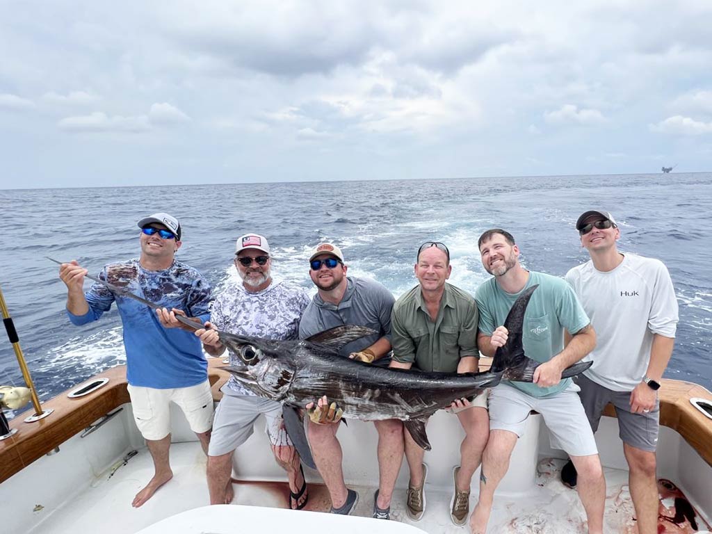 A group of six male anglers sitting on the deck of a fishing boat in the Gulf of Mexico on a cloudy day, holding a large swordfish