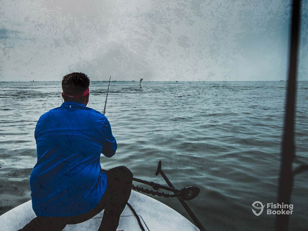 A view from behind of an angler fishing off the front of a fishing charter in the inshore waters of Crystal River, with a Tarpon visible at the end of the line in the distance, leaping out of the water on a cloudy day