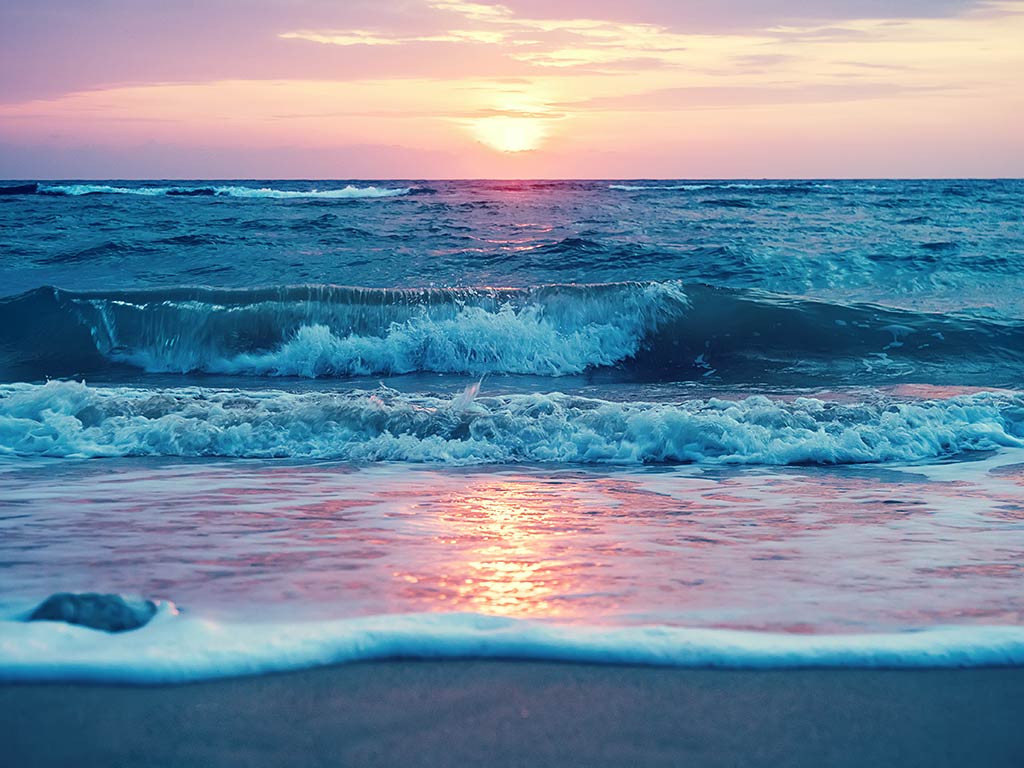 A view from the beach of waves crashing into it in Hawaii, with the sun setting in the distance