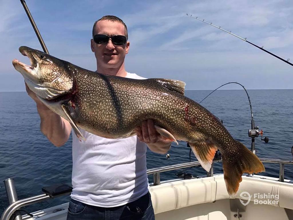 An angler with short hair and wearing sunglasses holds a Large Lake Trout aboard a fishing charter in Illinois, with clear blue waters visible behind him on a sunny day