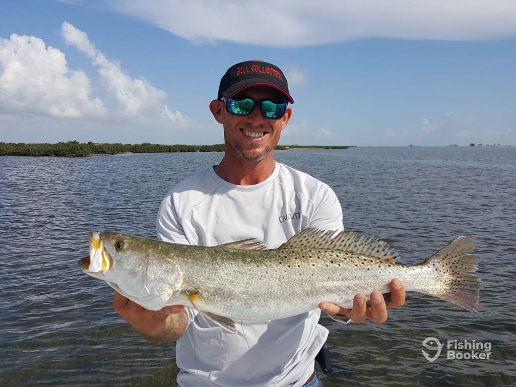 An angler in a baseball cap and sunglasses holding a large Speckled Trout in front of his chest on a sunny day in Port Isabel, with the water behind him and a shoreline visible in the distance