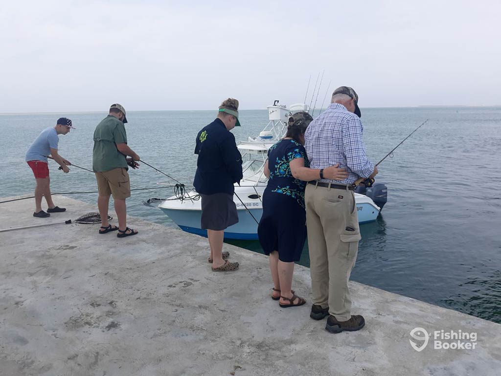 A group of anglers on a dock in Wanchese, with two pulling a boat towards the dock by a rope, two looking at the water, while the one to the furthest on the right casts a fishing rod into the water on a cloudy day