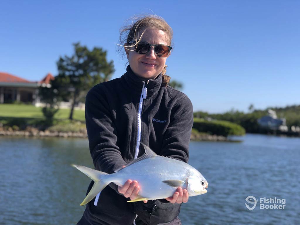 A woman wearing sunglasses, holding a Pompano caught in calm inshore waters with some land visible behind her on a sunny day