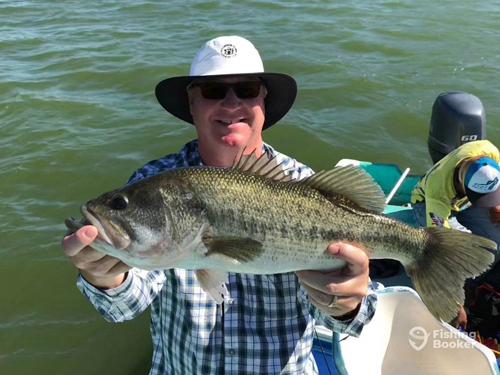 An angler in a hat posing with a Bass caught fishing in Mexico with the water visible behind him