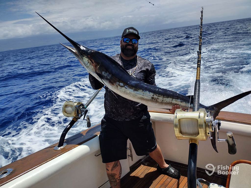 A bearded angler wearing a hat holds a large Marlin on the deck of an offshore sportfishing boat, with the wake of the boat visible in the water behind him and a reel of a trolling rod visible in the foreground