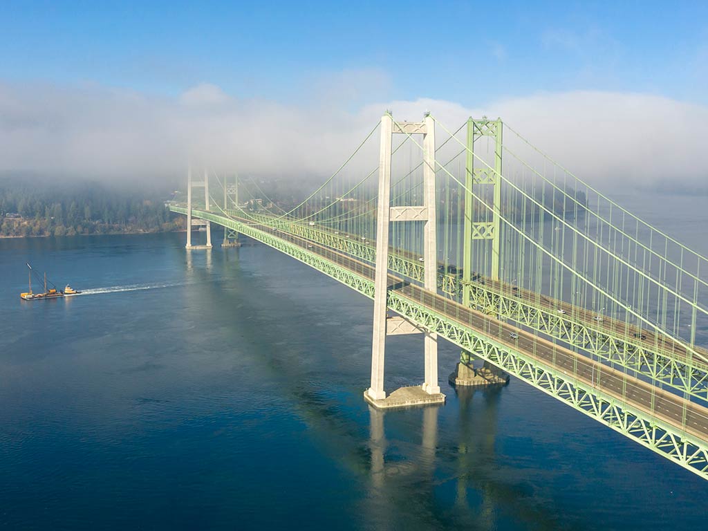 An aerial view of the Tacoma Narrows Bridge on a hazy day, with clouds visible in the distance obscuring part of the landscape