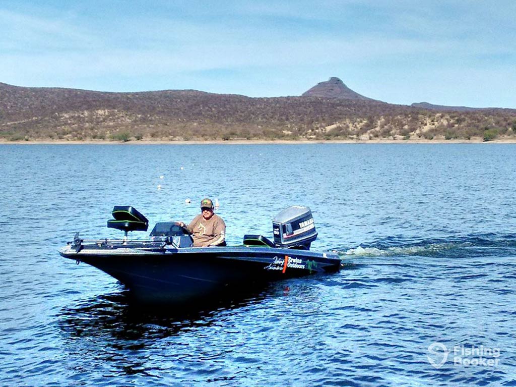 A view across the water towards a lone captain aboard his Bass fishing charter in Mexico, with a rugged landscape in the distance, including a pointed peak on a sunny day