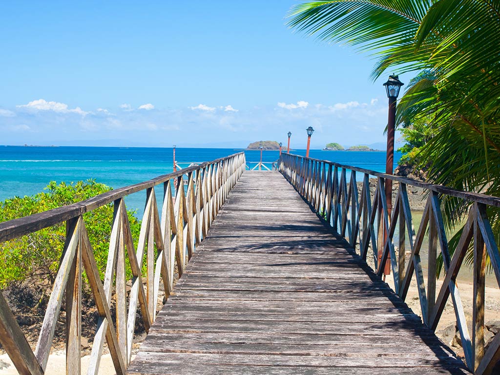 A view along a boardwalk towards the sea in Chiquirí, Panamá, on a clear day, with the turquoise waters of the ocean visible in the distance