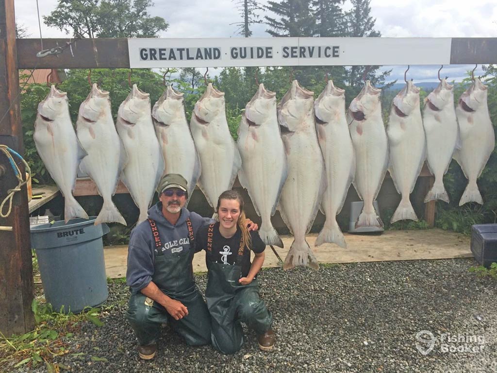 A father and daughter crouch down and pose in front of a sign saying "Greatland Guide Services", from which a number of Halibut are hanging