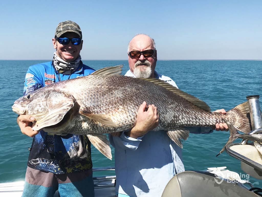 An elderly and younger angler hold a large Black Jewfish aboard a fishing charter in the Northern Territory on a sunny day with the blue waters of the ocean behind them