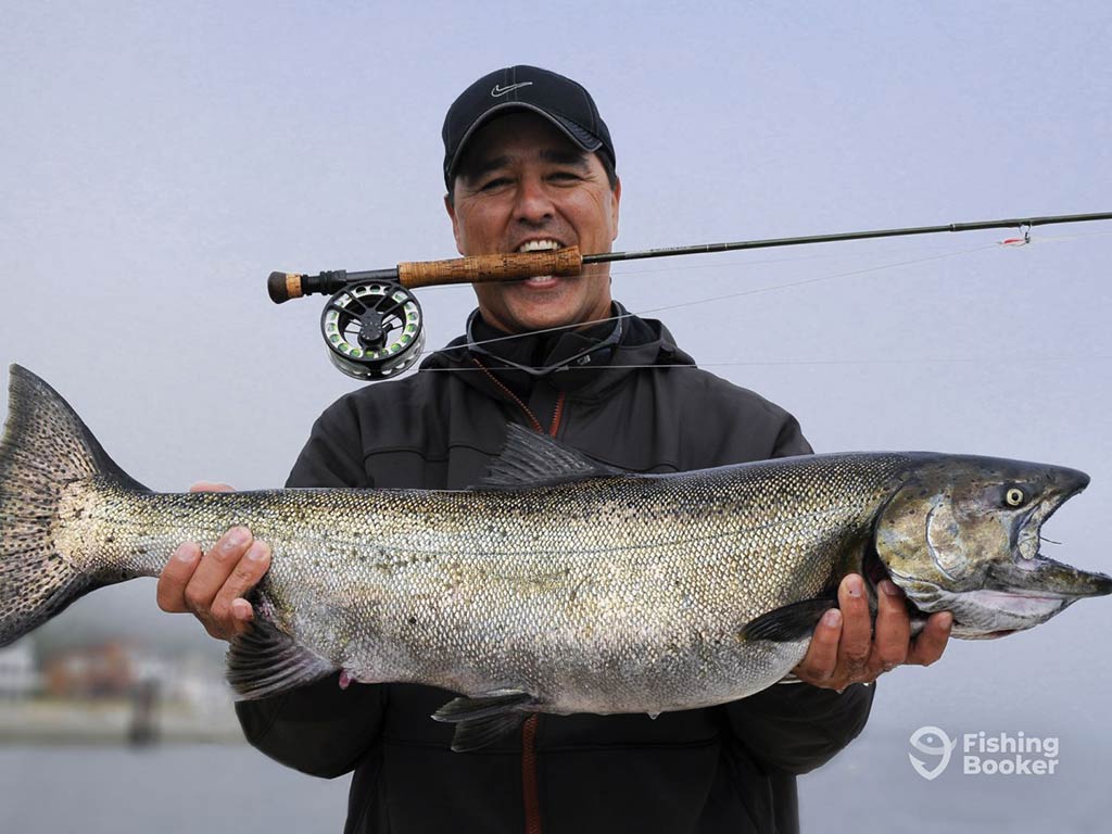 An angler holds a large Chinook Salmon while posing with his fly fishing rod in his mouth late on the day, with the water behind him