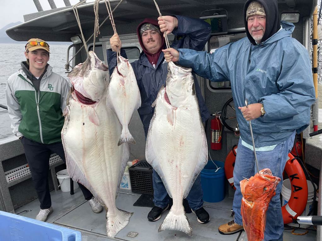 Two men in their twenties and one older man, standing on the deck of a fishing charter in Alaska, holding up two large Halibuts, one smaller, and a Rockfish on a cloudy day