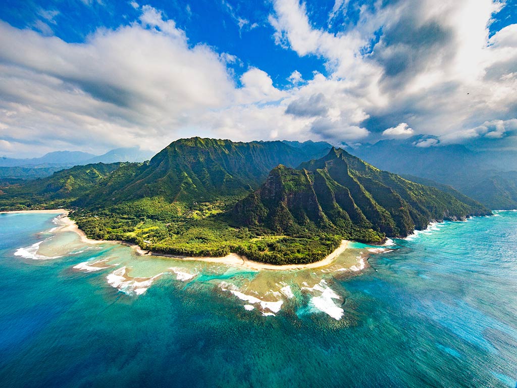 An aerial view of a a luscious, green, volcanic island in Hawaii, with turquoise waters and sandy beaches in the foreground on a sunny day