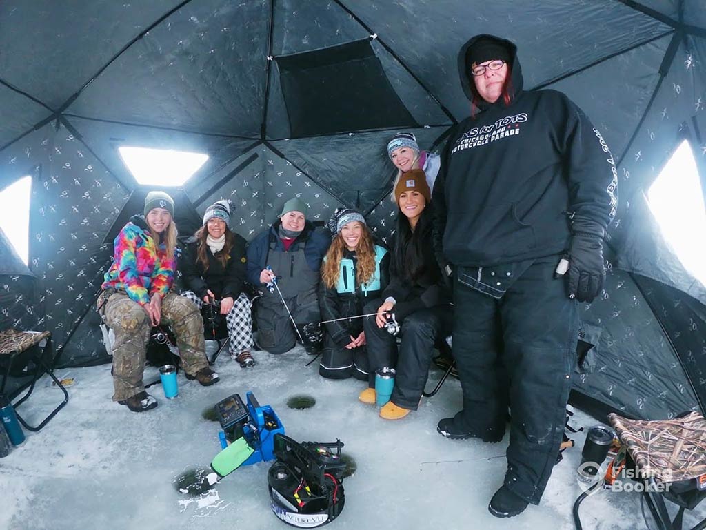 A group of female anglers crowded in an ice hut on a lake in Illinois, with ice fishing gear set up in front of them