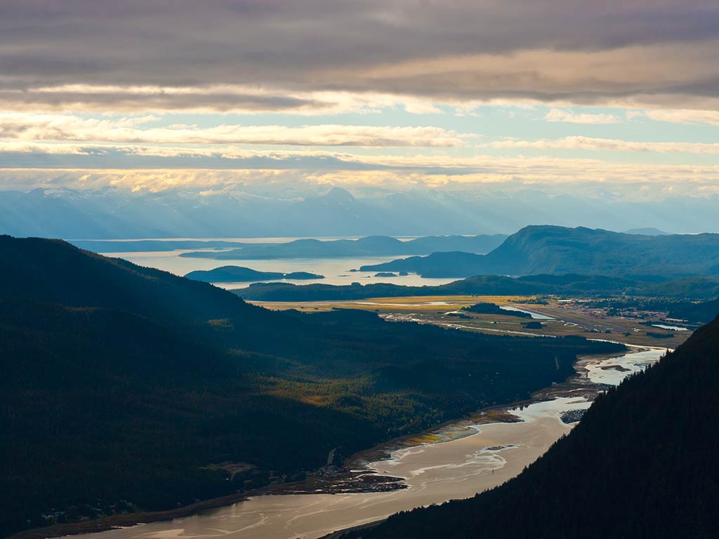 An aerial view of Juneau, Alaska, with a winding river dominating the centre of the image, making its way towards the ocean between two mountains on a cloudy day