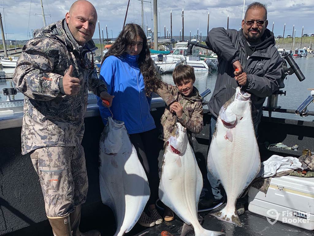 Two men, a boy, and girl standing on a fishing charter back at the dock in Homer, Alaska, holding up three Halibut between them, with docked boats visible behind them