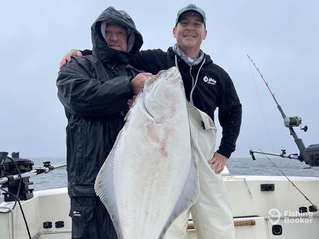 Two anglers, one wrapped up in winter gear and his face barely visible, holding a large Halibut caught in Alaska on a cloudy day with the water and a fishing rod visible behind them