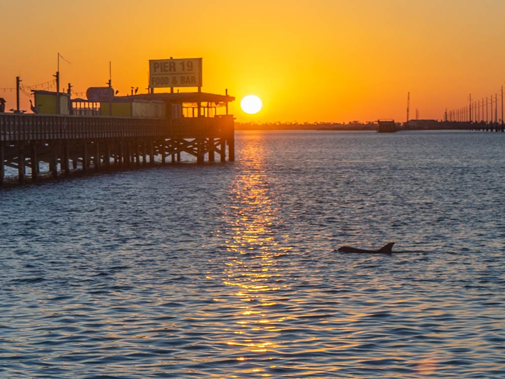 A view from a beach towards a fishing pier in South Padre Island, Texas at sunset, with the bright sun visible in the distance creating an orange hue on the horizon