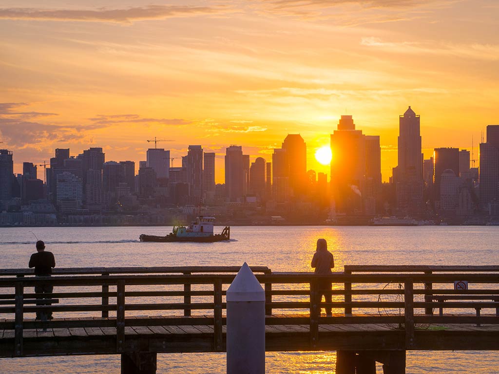 A view across a fishing pier towards the skyline of Seattle, with a silhouette of a fishing pier and a boat visible in the foreground, as the sun sets in the distance