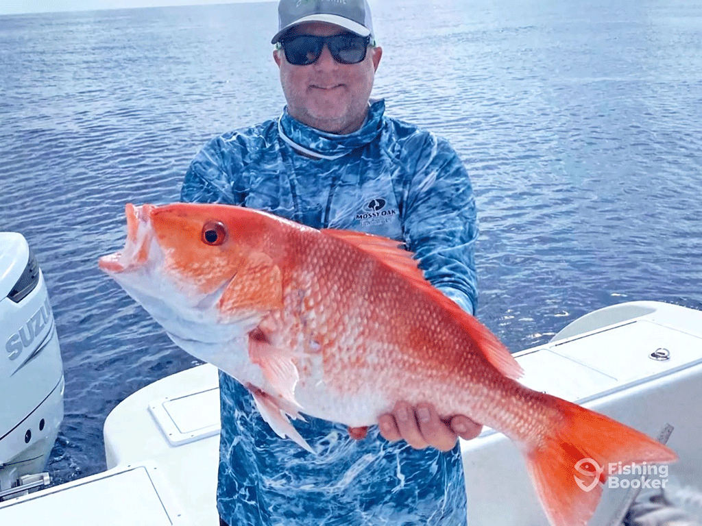 An angler in a camouflage blue shirt, sunglasses, and sunglasses holds a large Red Snapper aboard a Port Isabel offshore fishing charter on a sunny day, with the deep waters visible behind him