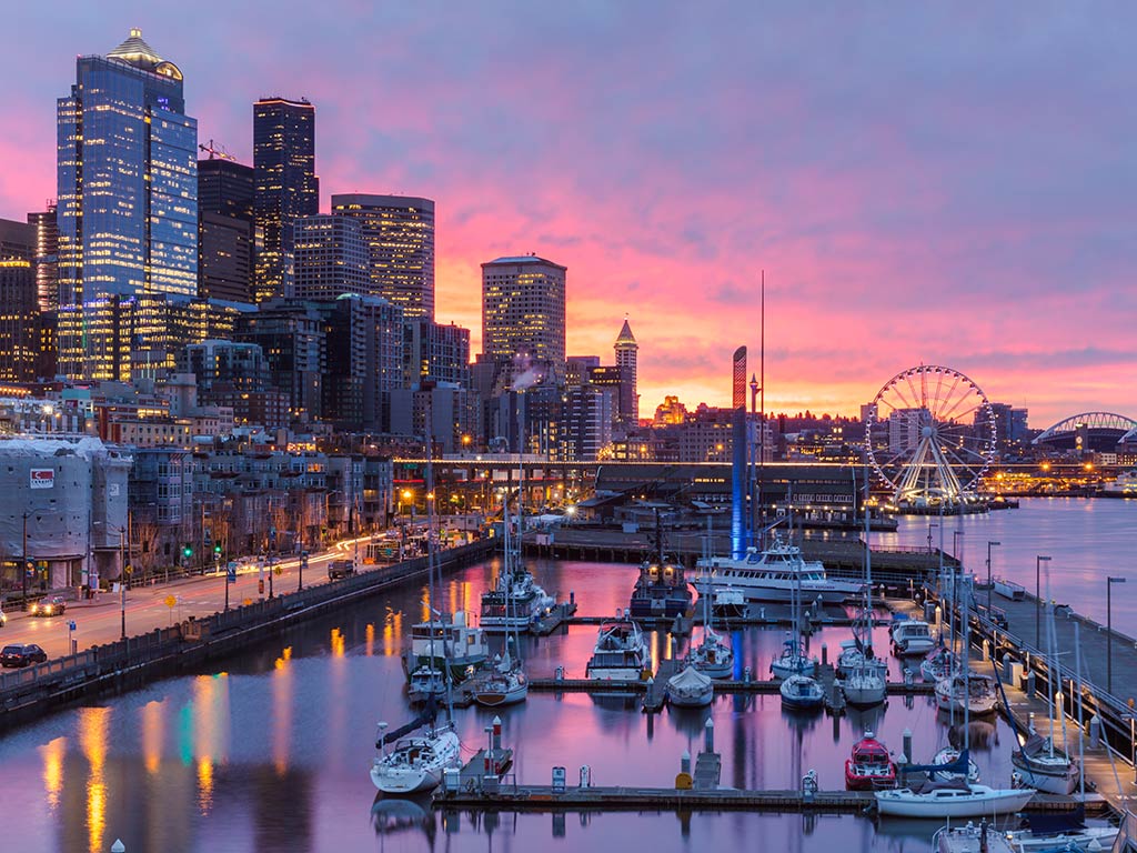 A view towards the skyline of Seattle from a marina, with boats visible in the foreground, the waters of Elliot Bay on the right of the image, and the sun setting in the distance behind the city's Ferris wheel, creating a purple hue in the sky