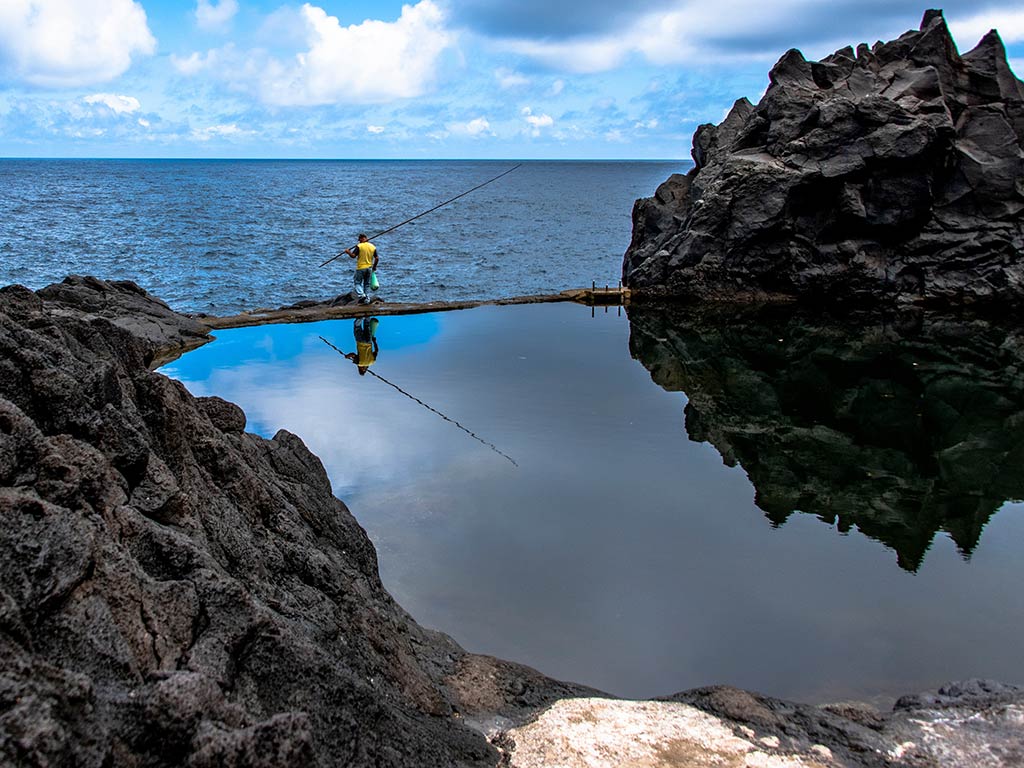 A view from behind of a distant angler casting from a calm pool of water towards the open ocean along Madeira's rocky coastline