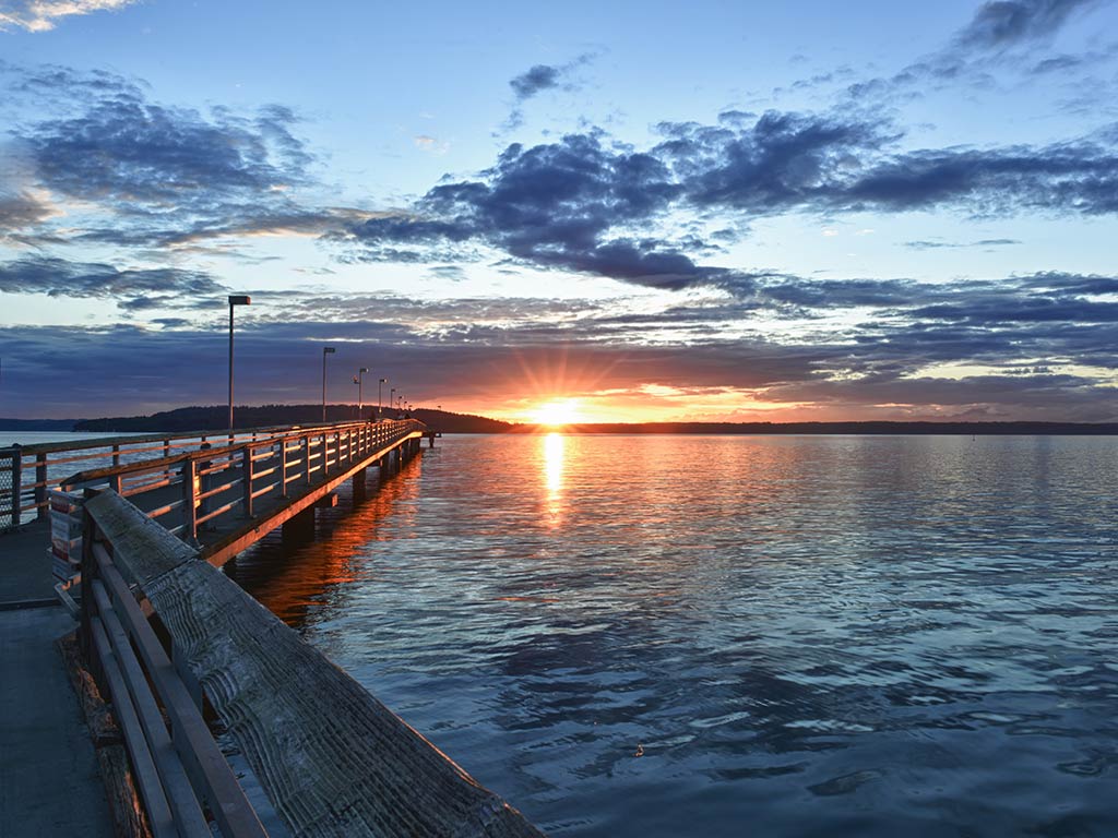 A view across a fishing pier in Puget Sound towards the sun setting amid low-lying clouds at the end of a relatively clear day