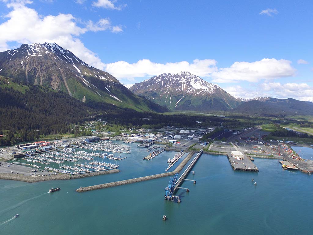 An aerial view looking toward Seward, AK's harbor on a day with sunny intervals, with a couple of boats making their way back into the harbor visible on the water, a packed marina, and snow-capped mountains visible in the distance