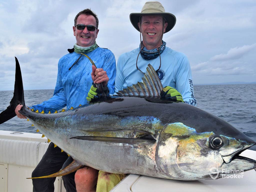 Two anglers – one wearing a straw hat – sitting on the side of a boat in Panamá and holding a large Tuna on a cloudy day