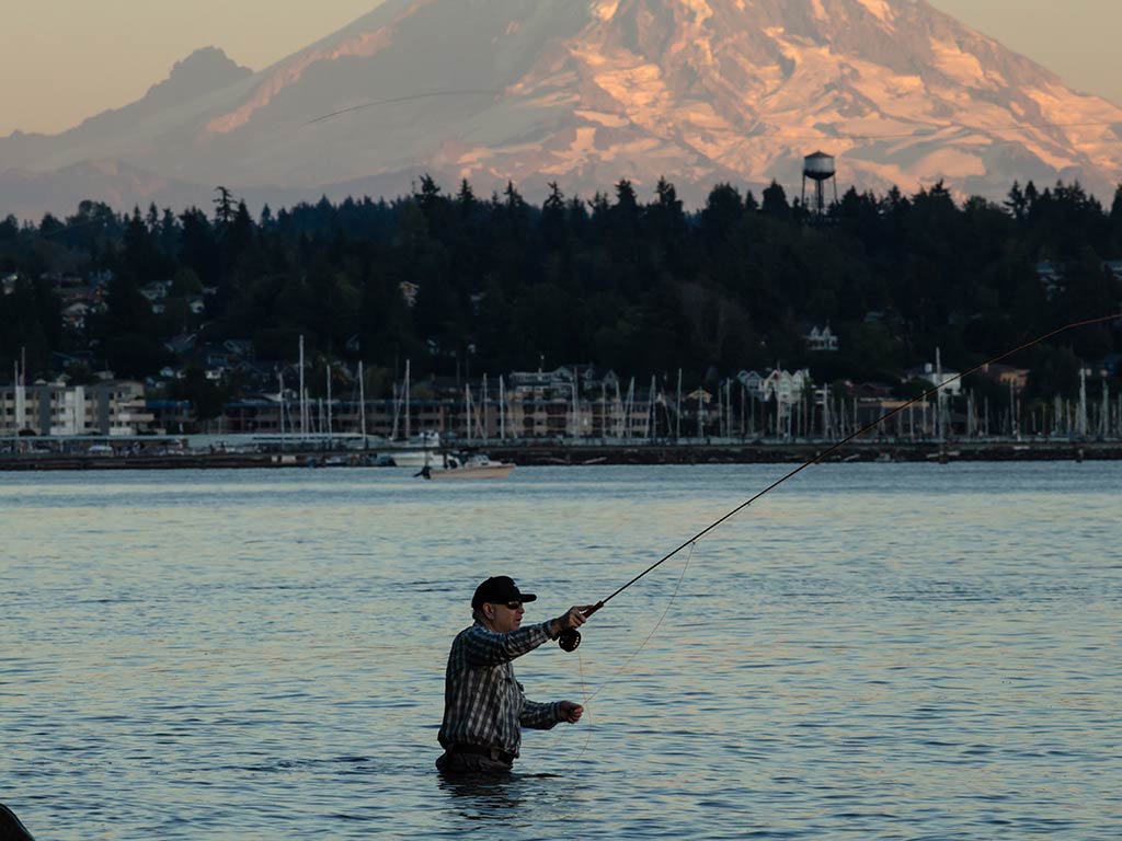 An angler up to his waist casts a line in the waters of Puget Sound at sunset against the backdrop of a snow-capped Mount Rainier