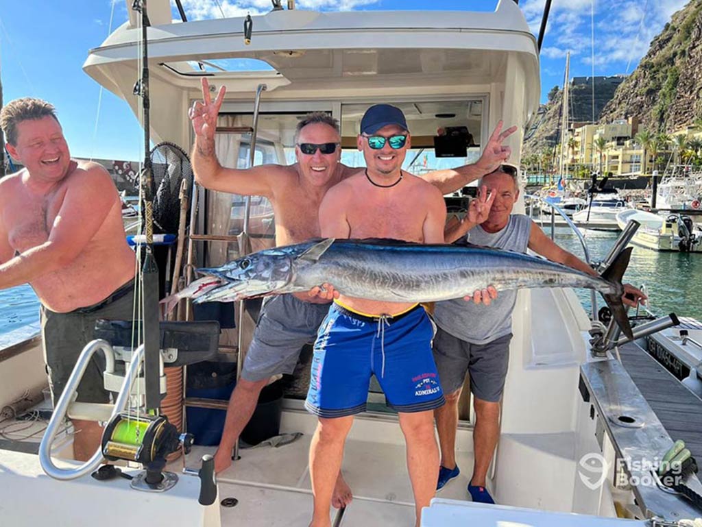 A shirtless angler standing on the decl of a fishing boat, back at the dock in Madeira, holds a large Wahoo, while his friends look on and smile