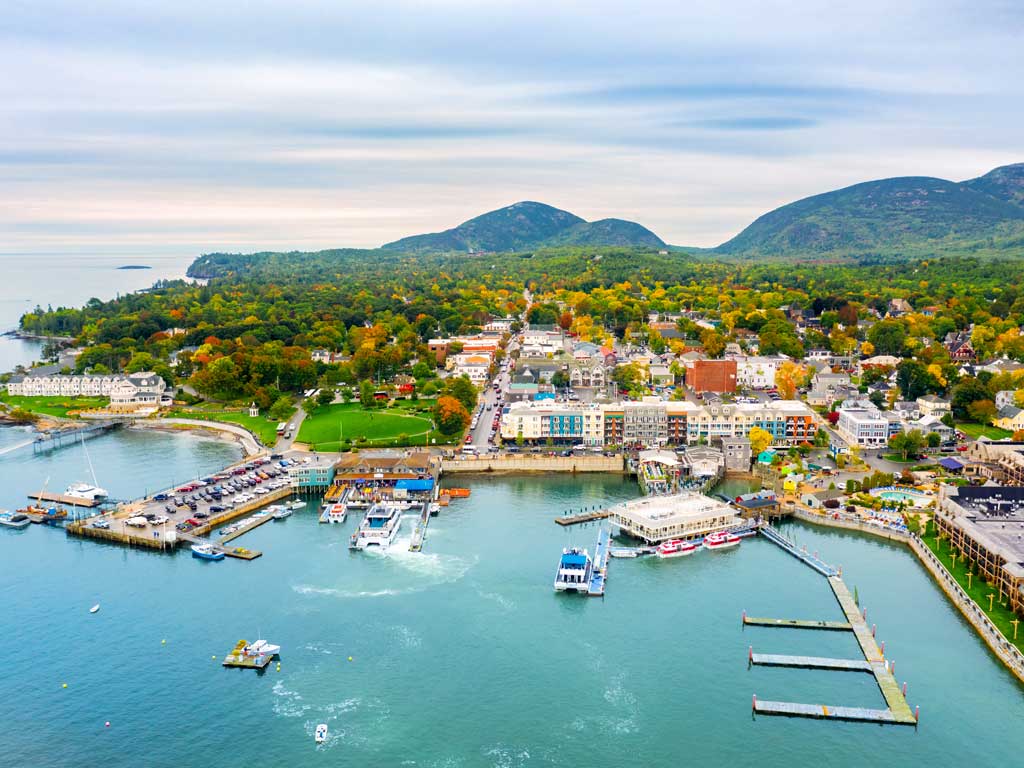 An aerial view of a bay dotted with charter fishing boats and marina, a town, its buildings, roads, cars, greenery, and several mountains in the background in Bar Harbor in Maine