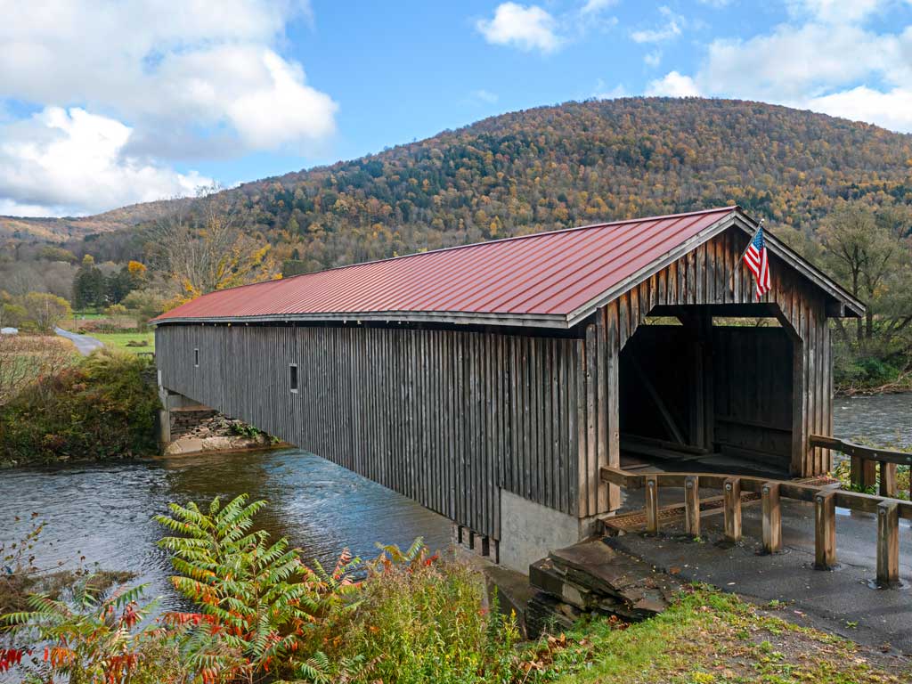 A photo of a roofed wooden bridge on the West Branch of the Delaware River near Hamden in the Great Western Catskills in New York State on a fall day