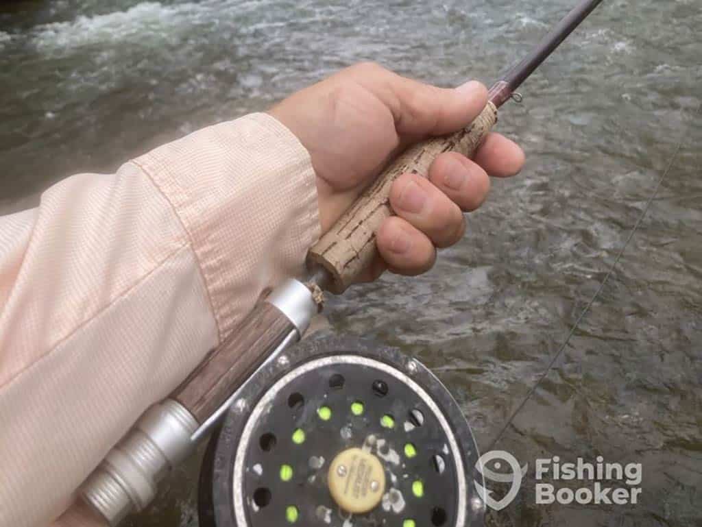 A closeup of a hand holding a fly fishing rod, with the reel visible in the foreground and the FishingBooker logo in the bottom right of the image